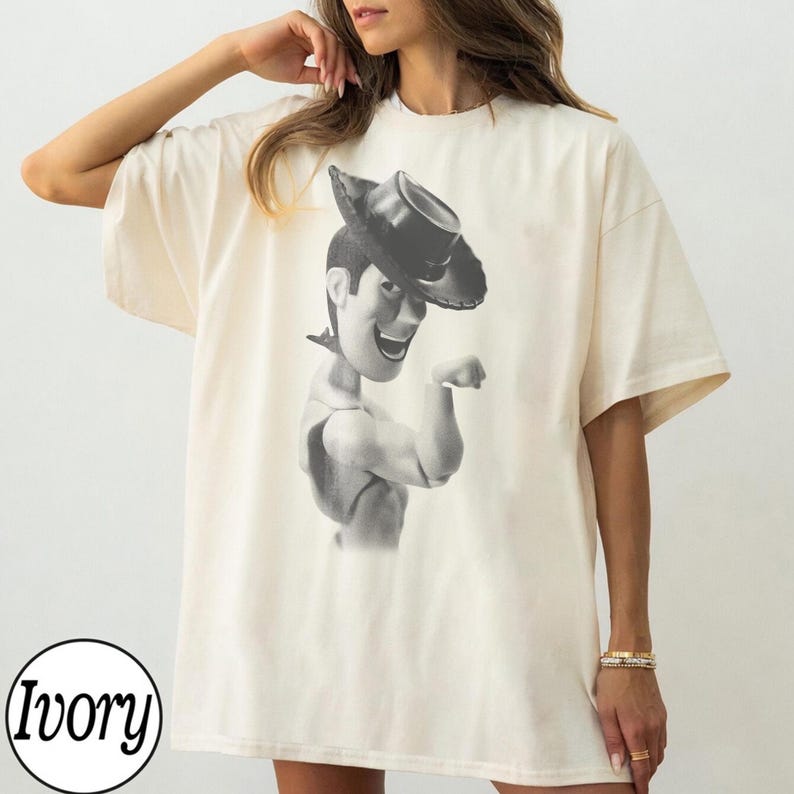 Puede incluir: Una camiseta de manga corta color marfil con un estampado gr&aacute;fico en blanco y negro de un personaje vaquero con un sombrero de ala ancha. El dise&ntilde;o est&aacute; centrado en el pecho y muestra al personaje desde el torso hacia arriba sonriendo.