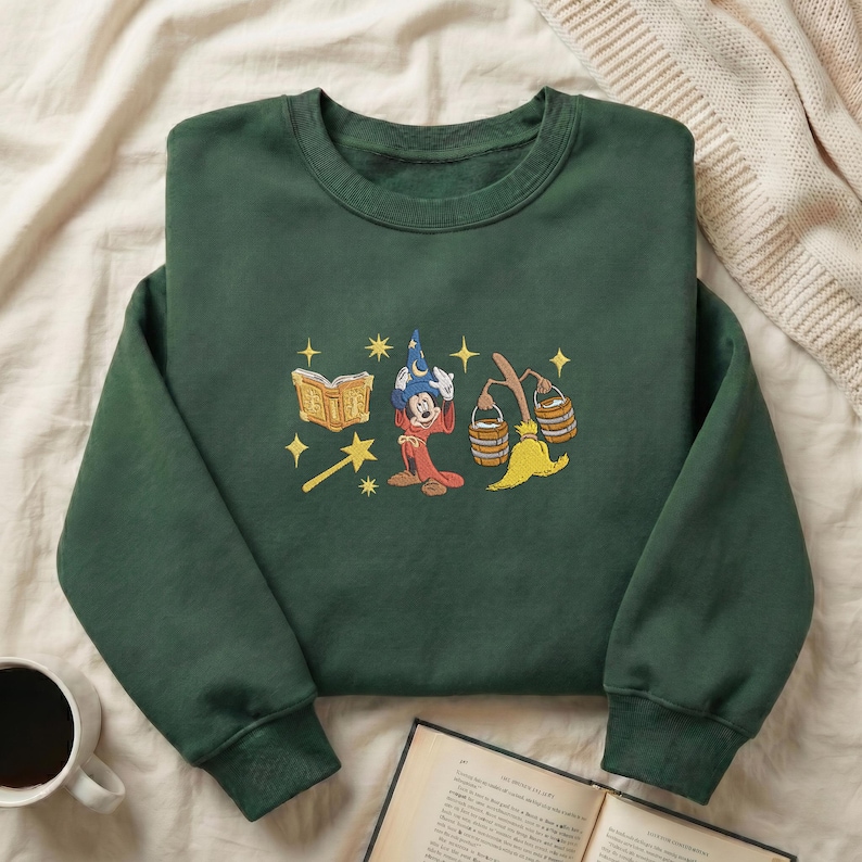 May include: Dark green sweatshirt with an embroidered design of a wizard, book, stars, and a broom. The design includes a wizard in a red robe and blue hat. The sweatshirt is made of a soft material and is perfect for fans of fantasy.