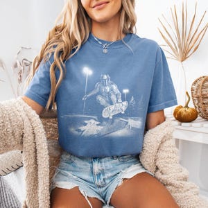 May include: A blue short-sleeve t-shirt featuring a white graphic print of a character in armor riding a skateboard in a skate park at night, with a small creature figure below. The shirt has a crew neckline and a relaxed, casual fit.
