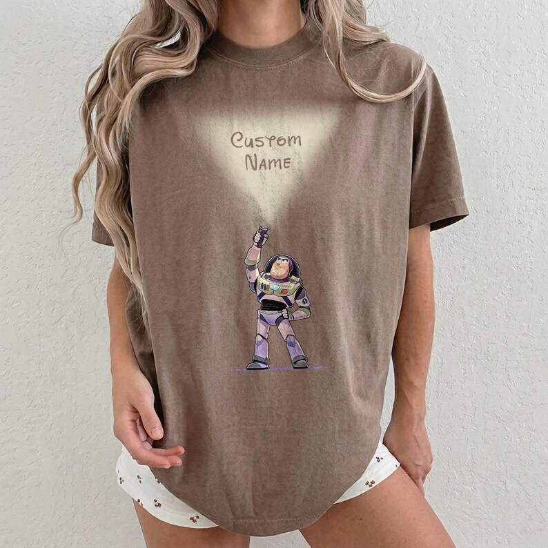 Pu&ograve; includere: T-shirt marrone con un'illustrazione di Buzz Lightyear di Toy Story. Il personaggio &egrave; raffigurato nella sua tuta spaziale, con la scritta "Custom Name" sopra. La maglietta &egrave; di colore neutro, adatta all'uso quotidiano.