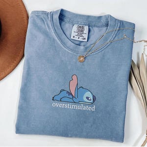 May include: A blue, pigment-dyed t-shirt featuring a graphic of the character Stitch lying down with the text "overstimulated" printed underneath. The shirt has a crew neckline and a Comfort Colors brand label visible at the inner collar.