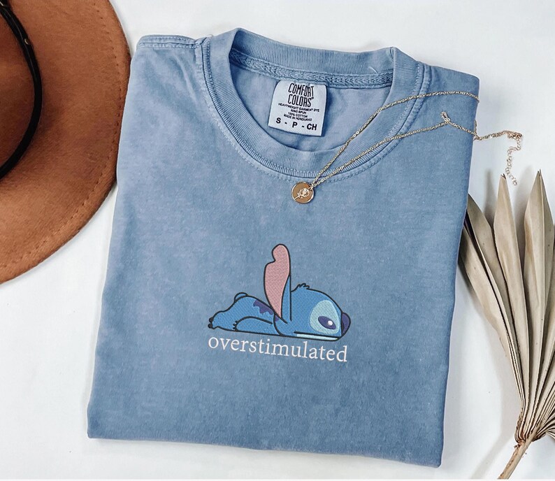 May include: A blue, pigment-dyed t-shirt featuring a graphic of the character Stitch lying down with the text "overstimulated" printed underneath. The shirt has a crew neckline and a Comfort Colors brand label visible at the inner collar.