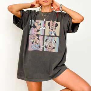 May include: Dark gray oversized t-shirt featuring a four-panel graphic of Minnie Mouse in various poses. Each panel has a pastel background. Minnie is wearing different bows and outfits. The shirt has short sleeves and a relaxed fit.