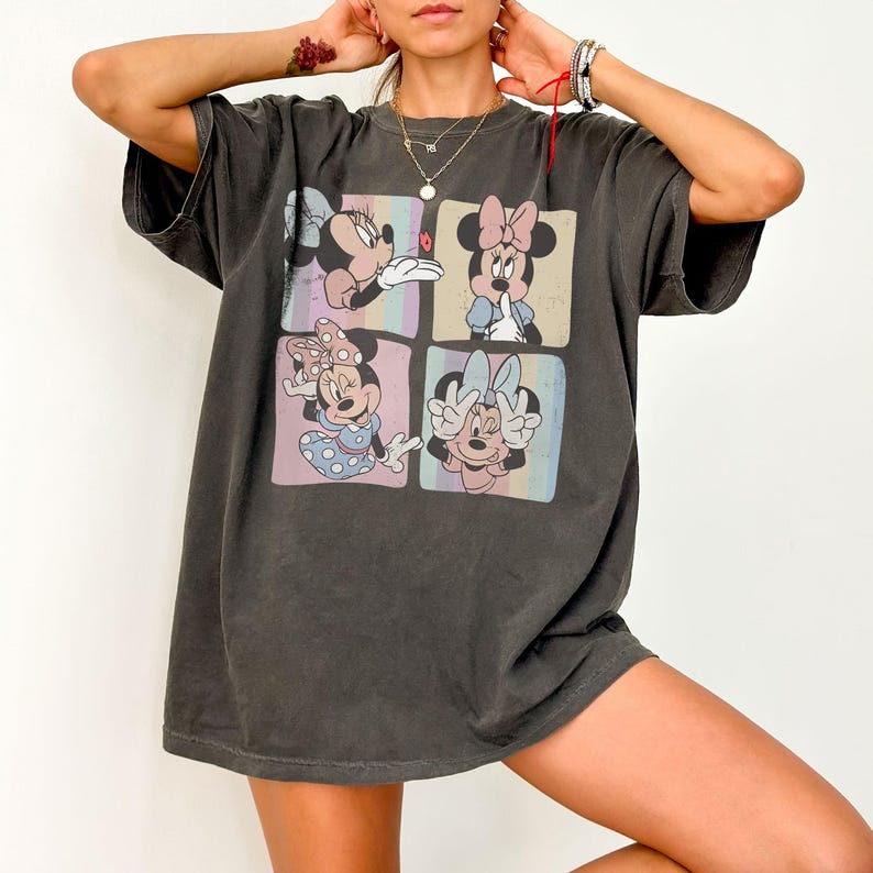 May include: Dark gray oversized t-shirt featuring a four-panel graphic of Minnie Mouse in various poses. Each panel has a pastel background. Minnie is wearing different bows and outfits. The shirt has short sleeves and a relaxed fit.