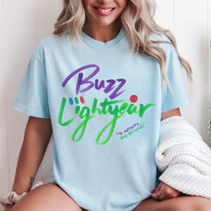 Puede incluir: Una camiseta de manga corta de color azul claro con el texto morado Buzz y el texto verde Lightyear. Sobre la palabra Lightyear hay peque&ntilde;os puntos rojos, azules y verdes. Debajo aparece el texto verde To Infinity... And Beyond! en cursiva.