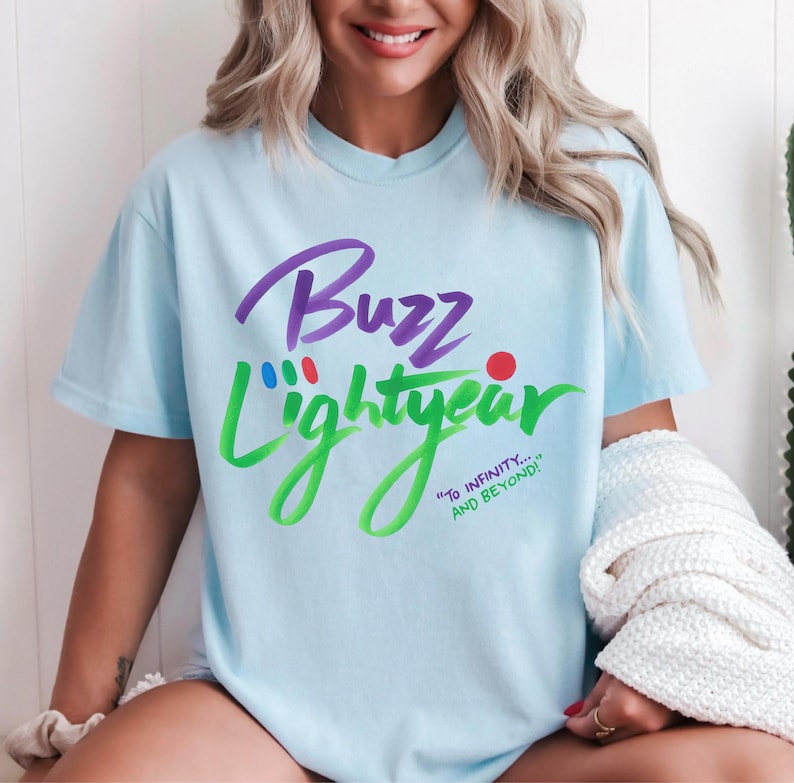 Puede incluir: Una camiseta de manga corta de color azul claro con el texto morado Buzz y el texto verde Lightyear. Sobre la palabra Lightyear hay peque&ntilde;os puntos rojos, azules y verdes. Debajo aparece el texto verde To Infinity... And Beyond! en cursiva.
