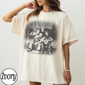 May include: An ivory short-sleeved t-shirt featuring a black and white graphic print of the characters Buzz Lightyear and Woody sitting together. The design has a faded, vintage-style aesthetic with a soft-focus background of a suburban street.