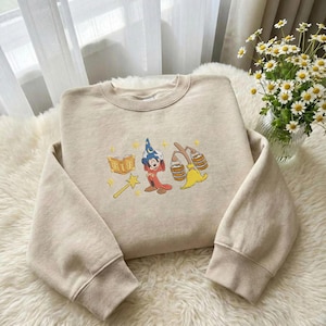 May include: Beige sweatshirt featuring an embroidered design of Mickey Mouse dressed as a wizard, a book, a magic wand, and a broom. The sweatshirt has a crew neck and long sleeves with ribbed cuffs. The design is in yellow, red, blue, and beige.