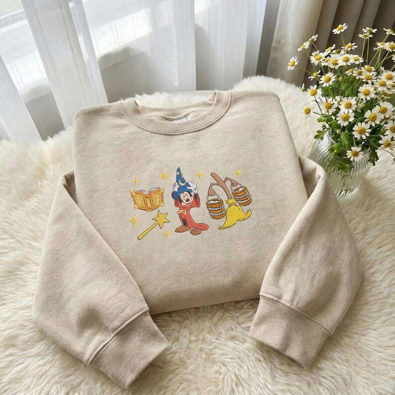 May include: Beige sweatshirt featuring an embroidered design of Mickey Mouse dressed as a wizard, a book, a magic wand, and a broom. The sweatshirt has a crew neck and long sleeves with ribbed cuffs. The design is in yellow, red, blue, and beige.