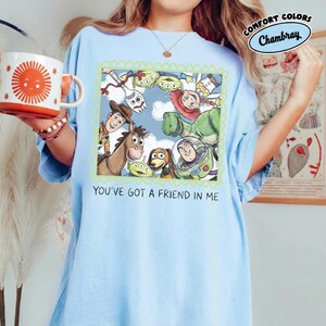 May include: Light blue t-shirt featuring a framed illustration of characters from the animated film "Toy Story". The image includes Woody, Buzz Lightyear, Rex, and other characters. The text "YOU'VE GOT A FRIEND IN ME" is printed below the illustration. The shirt has a relaxed fit.