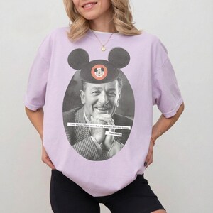 May include: Lilac-colored t-shirt featuring a black and white oval portrait of Walt Disney wearing a Mickey Mouse hat. The shirt includes the quote, "I love Mickey Mouse more than any woman I have ever known."