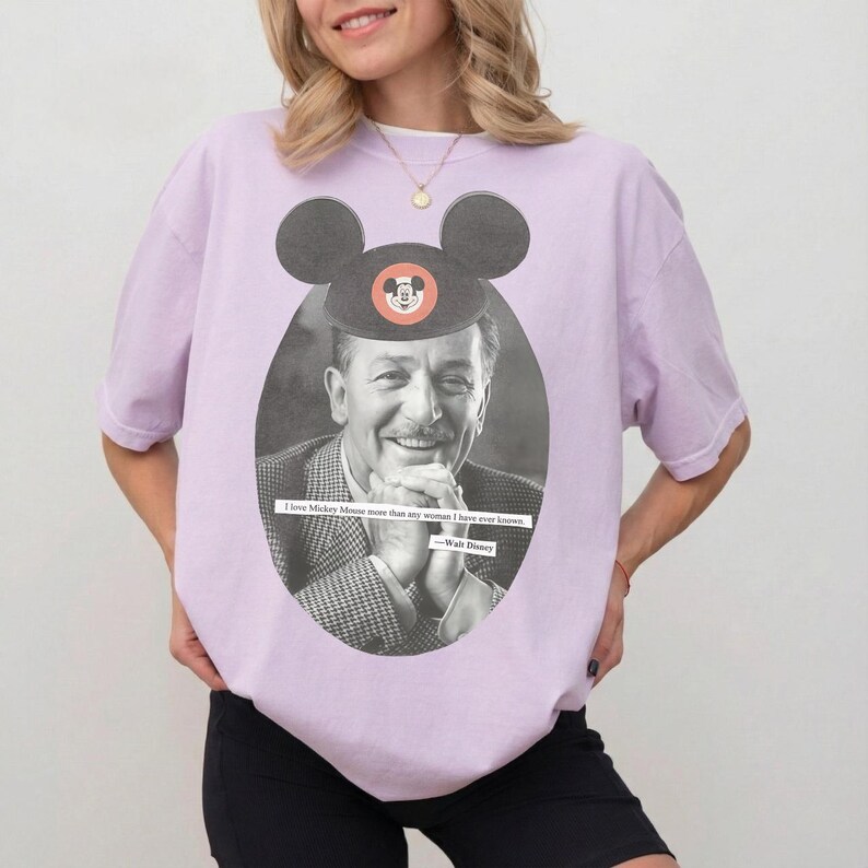 May include: Lilac-colored t-shirt featuring a black and white oval portrait of Walt Disney wearing a Mickey Mouse hat. The shirt includes the quote, "I love Mickey Mouse more than any woman I have ever known."
