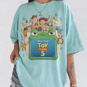 Może przedstawiać: Jasnoniebieski t-shirt z nadrukiem graficznym przedstawiającym r&oacute;żne postacie z Toy Story, w tym Chudego, Jessie i Buzza Astrala. Pośrodku znajduje się zielona ramka w kształcie żaby z napisem Disney Pixar Toy Story 5 oraz nutami muzycznymi.