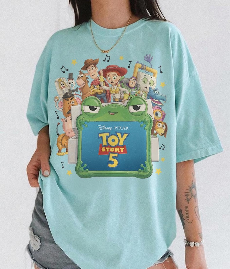 Może przedstawiać: Jasnoniebieski t-shirt z nadrukiem graficznym przedstawiającym r&oacute;żne postacie z Toy Story, w tym Chudego, Jessie i Buzza Astrala. Pośrodku znajduje się zielona ramka w kształcie żaby z napisem Disney Pixar Toy Story 5 oraz nutami muzycznymi.