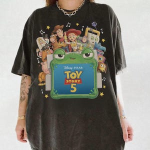 Może przedstawiać: Czarny t-shirt z kr&oacute;tkim rękawem z nadrukiem graficznym przedstawiającym r&oacute;żne postacie z Toy Story otaczające dużą zieloną ramkę w kształcie żaby. Wewnątrz ramki znajduje się napis Disney Pixar Toy Story 5 oraz ż&oacute;łte gwiazdki i nuty.