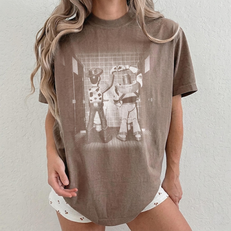 May include: A brown short-sleeve t-shirt featuring a monochromatic graphic print of two characters standing at urinals in a tiled restroom. The design depicts a cowboy figure on the left and a space ranger figure on the right, both facing away.