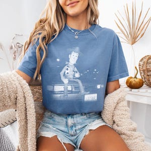 May include: A blue crew neck t-shirt featuring a graphic print of the character Woody from Toy Story sitting on the hood of a car. The graphic is rendered in a monochromatic white tone, showing the character, the car headlights, and a license plate.