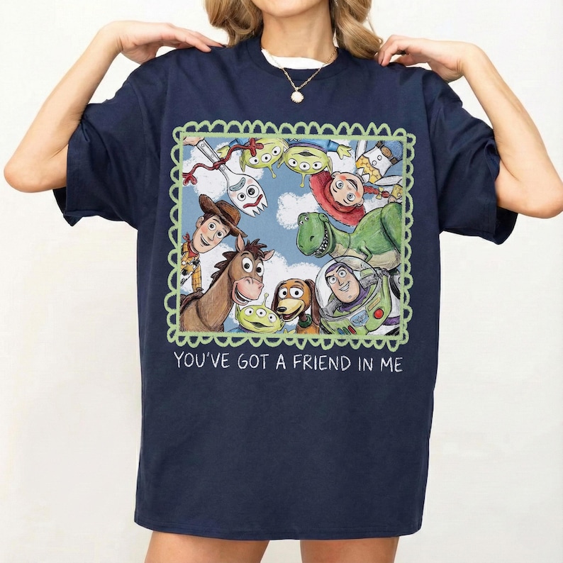 May include: Navy blue t-shirt featuring a colorful illustration of characters from the movie Toy Story, including Woody, Buzz Lightyear, and others, within a green scalloped frame. The text "YOU'VE GOT A FRIEND IN ME" is printed below the image.