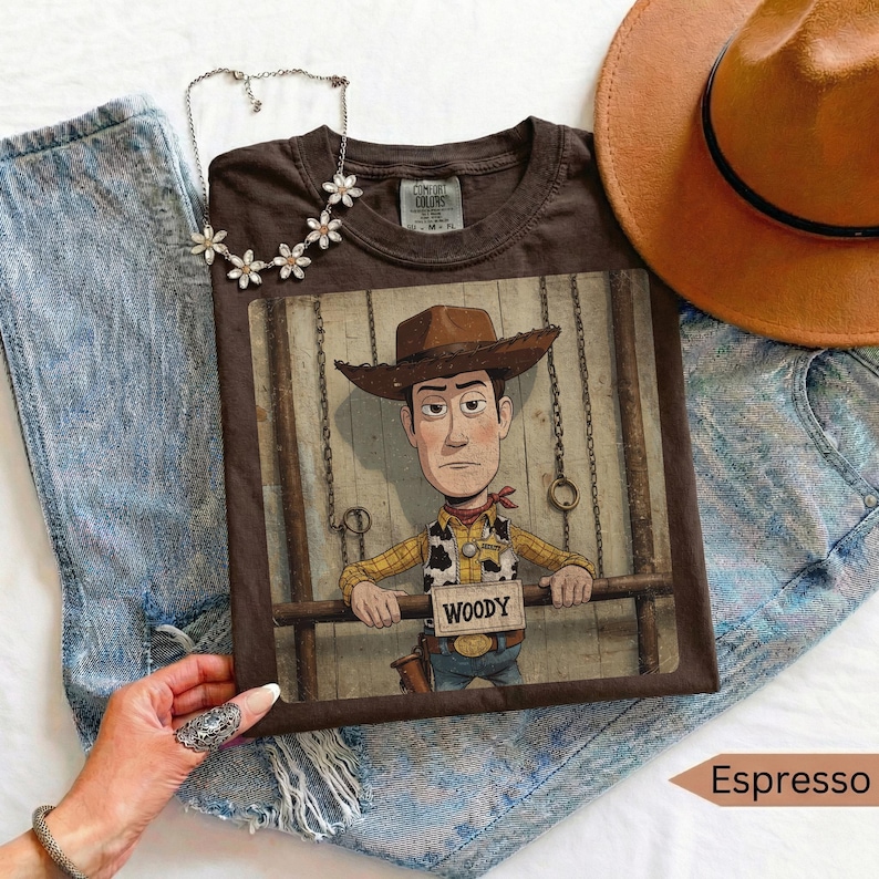 May include: A dark brown t-shirt featuring a graphic of the character Woody from Toy Story, who is wearing a cowboy hat and a yellow shirt with a vest. The character is positioned behind a wooden fence. A silver flower-shaped necklace is placed above.