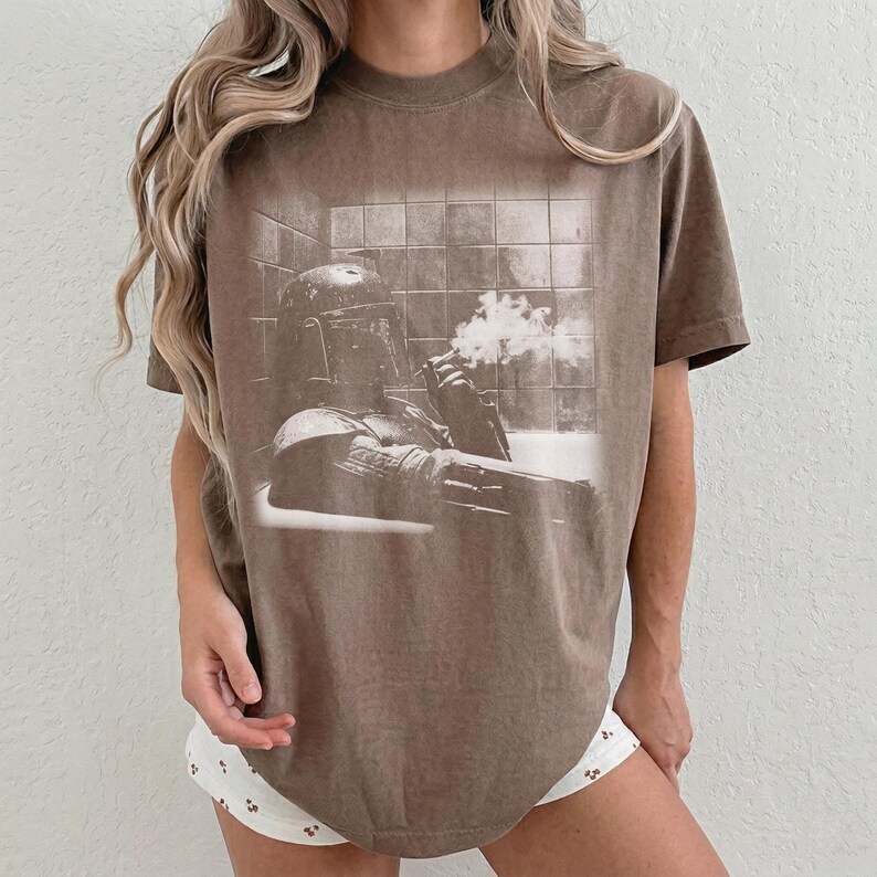 May include: A brown short-sleeve t-shirt featuring a monochromatic graphic print of a helmeted character sitting in a bathtub, holding a smoking object. The design shows the character in a tiled bathroom setting with visible smoke plumes.