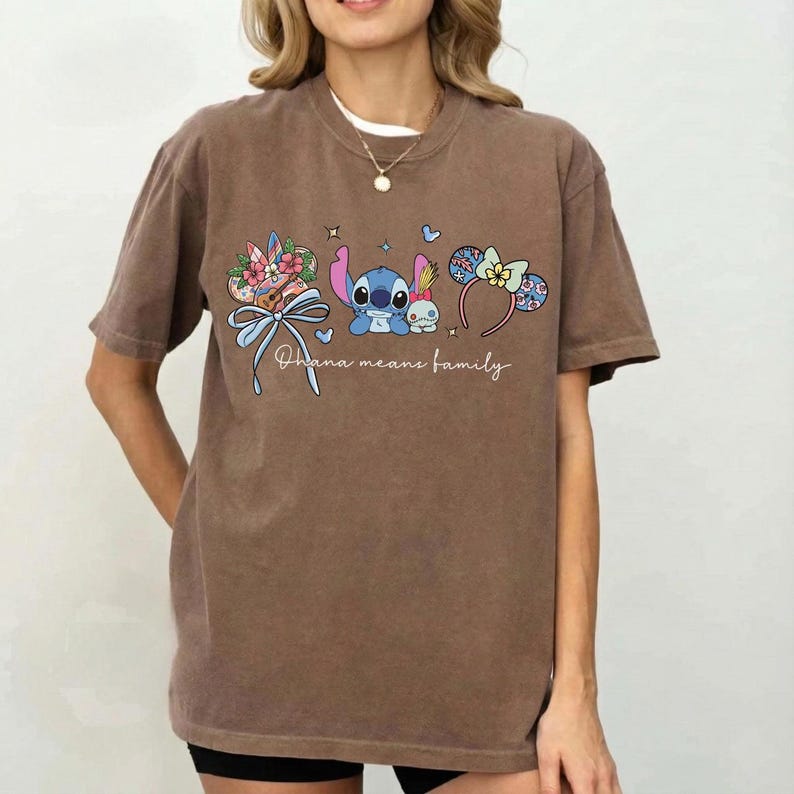 Puede incluir: Camiseta marr&oacute;n con un gr&aacute;fico de Stitch y Angel, detalles florales y lazos, y el texto escrito "Ohana means family". La camiseta tiene mangas cortas y cuello redondo.
