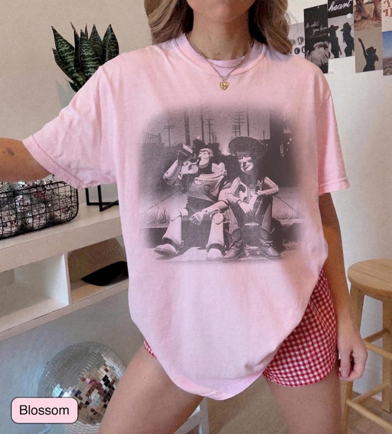 May include: A light pink short-sleeve t-shirt featuring a black and white graphic print of two animated characters sitting together on a curb. The shirt has a crew neckline and a loose, relaxed fit, showcasing a vintage-style screen-printed design.