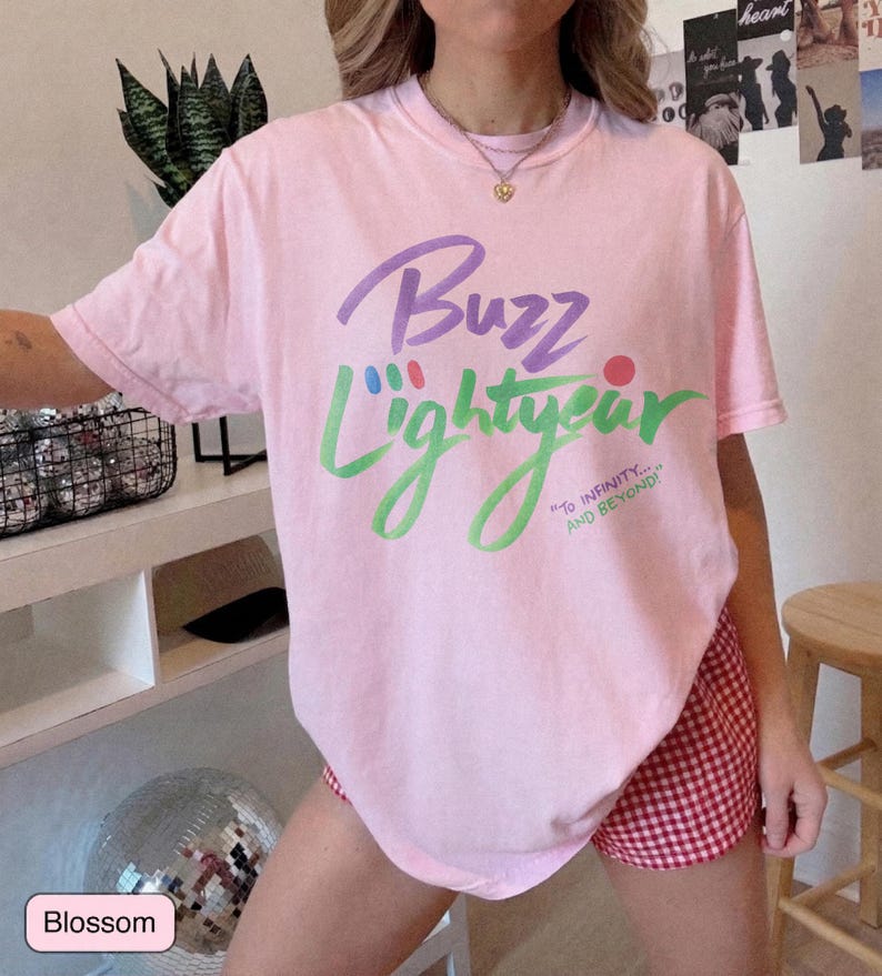 Puede incluir: Una camiseta de manga corta de color rosa claro con el texto Buzz Lightyear en morado y verde, junto con la frase To Infinity... and Beyond! impresa en verde. La camiseta se combina con unos pantalones cortos de cuadros rojos y blancos.