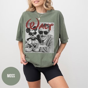 May include: An olive green t-shirt featuring a black and white graphic of Mickey Mouse wearing sunglasses and Walt Disney. The word "WALT" is printed in red script above the image. The shirt is a relaxed fit.