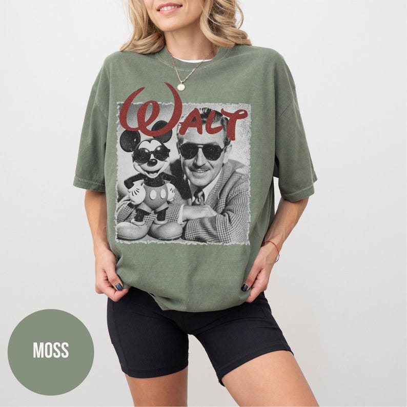 May include: An olive green t-shirt featuring a black and white graphic of Mickey Mouse wearing sunglasses and Walt Disney. The word "WALT" is printed in red script above the image. The shirt is a relaxed fit.