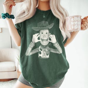 May include: A dark green short-sleeve t-shirt featuring a graphic print of a cowboy character wearing a hat, a vest with a sheriff badge, and a patterned shirt. The character is depicted with hands near the face and tongue sticking out.