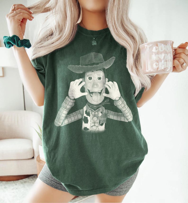 May include: A dark green short-sleeve t-shirt featuring a graphic print of a cowboy character wearing a hat, a vest with a sheriff badge, and a patterned shirt. The character is depicted with hands near the face and tongue sticking out.