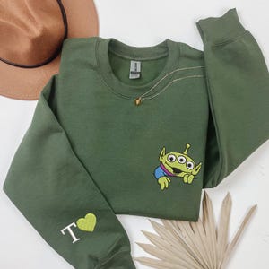 May include: An olive green crewneck sweatshirt featuring an embroidered three-eyed green alien character on the chest and a white letter T followed by a lime green heart embroidered on the left sleeve cuff, with a gold chain necklace.