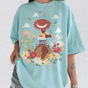 May include: A light blue t-shirt featuring a graphic illustration of a cowgirl figure with a red hat, sitting on a brown horse. The design includes cacti, various colorful flowers, small white clouds, and musical notes scattered around the central figure.