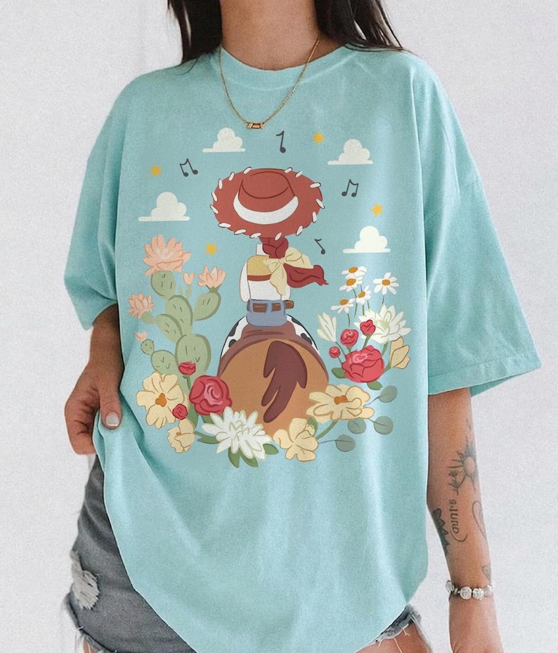 May include: A light blue t-shirt featuring a graphic illustration of a cowgirl figure with a red hat, sitting on a brown horse. The design includes cacti, various colorful flowers, small white clouds, and musical notes scattered around the central figure.
