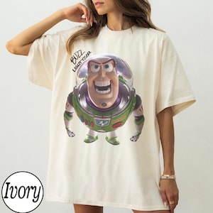 May include: An ivory short-sleeved t-shirt featuring a graphic print of the character Buzz Lightyear from Toy Story. The character is depicted in a space suit with a clear helmet. The text "BUZZ LIGHTYEAR" is printed in black above the character.