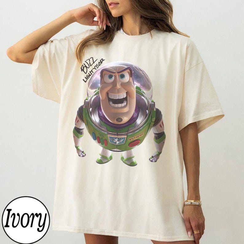 May include: An ivory short-sleeved t-shirt featuring a graphic print of the character Buzz Lightyear from Toy Story. The character is depicted in a space suit with a clear helmet. The text "BUZZ LIGHTYEAR" is printed in black above the character.