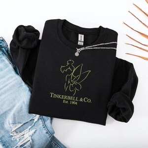 Pode incluir: Um moletom preto com gola redonda com um gr&aacute;fico verde lim&atilde;o de uma fada e o texto "Tinkerbell & Co. Est. 1904." Um colar de prata com um pingente circular est&aacute; no moletom. Jeans azuis claros rasgados &agrave; esquerda.