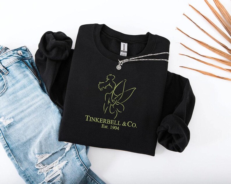 Pode incluir: Um moletom preto com gola redonda com um gr&aacute;fico verde lim&atilde;o de uma fada e o texto "Tinkerbell & Co. Est. 1904." Um colar de prata com um pingente circular est&aacute; no moletom. Jeans azuis claros rasgados &agrave; esquerda.