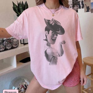 Puede incluir: Una camiseta de manga corta de color rosa claro con un estampado gr&aacute;fico en blanco y negro del personaje Woody de Toy Story con sombrero de vaquero. La camiseta tiene cuello redondo y un corte holgado, mostrando al personaje desde el pecho.