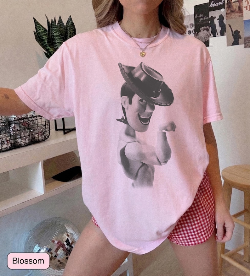 Puede incluir: Una camiseta de manga corta de color rosa claro con un estampado gr&aacute;fico en blanco y negro del personaje Woody de Toy Story con sombrero de vaquero. La camiseta tiene cuello redondo y un corte holgado, mostrando al personaje desde el pecho.