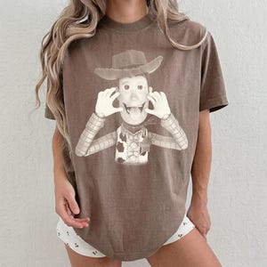 May include: A brown short-sleeve t-shirt featuring a monochromatic graphic print of a cowboy character wearing a hat, a vest with a cow-print pattern, and a sheriff badge. The character is depicted with hands near the face and tongue sticking out.