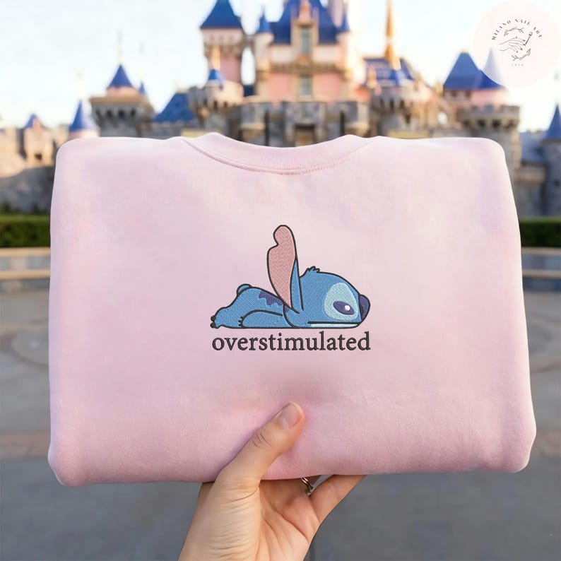 May include: A light pink crewneck sweatshirt featuring an embroidered graphic of a blue alien character lying down with its head resting on its arms. Below the character, the word "overstimulated" is embroidered in a dark brown lowercase font.