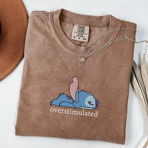 May include: A brown short-sleeved t-shirt featuring a graphic of a blue character lying down with the text "overstimulated" printed below. The shirt has a crew neckline and a Comfort Colors brand label on the inside collar showing size S-P-CH.