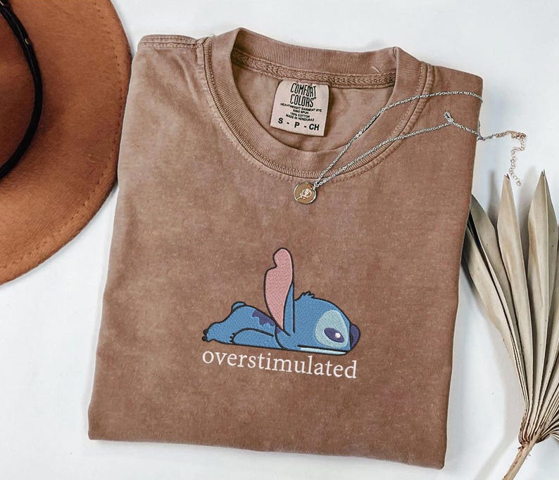 May include: A brown short-sleeved t-shirt featuring a graphic of a blue character lying down with the text "overstimulated" printed below. The shirt has a crew neckline and a Comfort Colors brand label on the inside collar showing size S-P-CH.