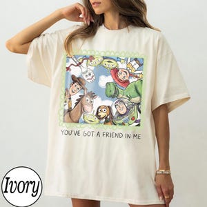 May include: Ivory-colored t-shirt featuring a colorful graphic of characters from the animated film "Toy Story." The graphic is framed with a green border and includes the text "You've Got a Friend in Me."