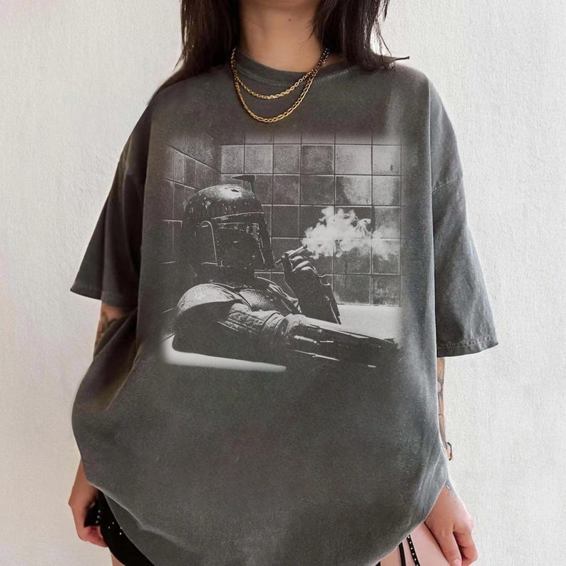 May include: A charcoal gray short-sleeve t-shirt featuring a black and white graphic print of a helmeted character in a bathtub with smoke rising from a cigarette. The shirt has a crew neckline and a relaxed fit, showcasing a vintage-style wash.