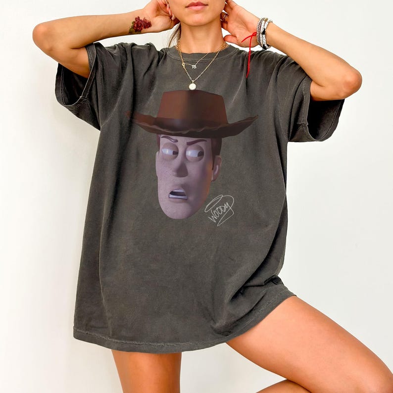 May include: A charcoal grey short-sleeve t-shirt featuring a graphic print of a cowboy character wearing a brown hat. The character has a wide-eyed, surprised expression. Below the character, the word Woody is written in a stylised white script.