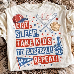 May include: Cream-colored t-shirt with a baseball theme. The shirt features a red and blue baseball cap, crossed bats, a baseball, and a glove. Text reads "EAT. SLEEP. TAKE KIDS TO BASEBALL! REPEAT!" with a "#1" foam finger.