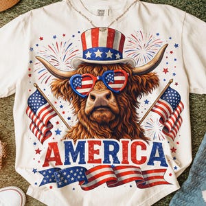 May include: White t-shirt featuring a Highland cow wearing heart-shaped American flag sunglasses and a patriotic top hat. The word "AMERICA" is displayed in red and blue banner. Two American flags and fireworks complete the design.