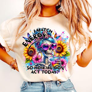 May include: A white t-shirt with a colorful graphic of a skeleton wearing sunglasses and a headband. The graphic includes the text "I Match Energy So How We Gon' Act Today?"